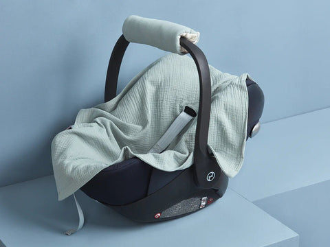 Ein schwarzer Kinderautositz mit einer hellgrünen Musselin-Decke darüber, vor einem minimalistischen blauen Hintergrund.