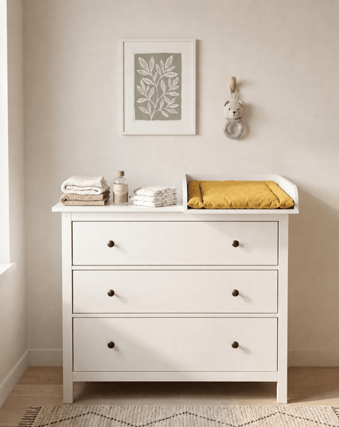 Eine weiße HEMNES Kommode mit einem KraftKids Wickelaufsatz (weiß, passend für 108 cm breite Kommode, Wickelfläche 50×70 cm) fasst Handtücher und eine gelbe Wickelauflage. Oben schmücken KraftKids Blattkunst und ein Tieraufhänger das sanft beleuchtete Zimmer.