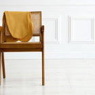 Ein Holzstuhl mit geflochtener Rohrrückenlehne steht auf einem hellen Boden. Über die Rückenlehne ist der KraftKids Stillkissenbezug Leinen gelb senfgelb drapiert. Weiß getäfelte Wände bilden den Hintergrund.