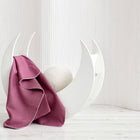 Ein weißer, halbmondförmiger Stuhl steht auf einem hellen Holzboden, drapiert mit der KraftKids Babydecke Leinen magenta dunkel und mit einem runden, strukturierten weißen Kissen. Im Hintergrund befinden sich weiße vertikale Jalousien.