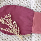 Ein KraftKids Strampelsack Frühling Sommer Leinen magenta dunkel ruht auf einer dicken weißen Strickdecke, die oben mit einem kleinen Zweig getrockneten Grases verziert ist.