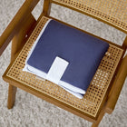 Eine gefaltete KraftKids Reisewickelunterlage Leinen blau dunkelblau mit weißem Gurt ruht auf der geflochtenen Sitzfläche eines Holzstuhls, der auf einem beigen Teppich steht.