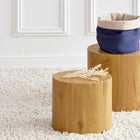 Zwei runde Holzhocker auf einem weißen, strukturierten Teppich vor einer weiß getäfelten Wand. Auf dem einen steht das KraftKids Körbchen rund Leinen blau dunkelblau, das sich perfekt für einen Wickeltisch eignet, auf dem anderen ein kleiner Strauß getrockneter beiger Blätter.