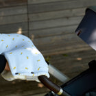 Die Hand einer Person, die von einem KraftKids Kinderwagenmuff Musselin mit gelben Zitronen warm gehalten wird, hält den Griff eines Kinderwagens im Freien an einer Holzwand.