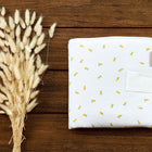 Eine gefaltete KraftKids Reisewickelunterlage Musselin gelbe Zitronen mit kleinen gelben Zitronenmustern liegt auf einer Holzfläche neben einem Bündel getrockneter, flauschiger beiger Grashalme.