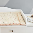 Ein weißer Wickeltisch mit einer weichen, pfirsichfarbenen KraftKids Wickelauflage Musselin Kirschen. Daneben liegt eine gefaltete weiße Decke mit einem kleinen Plüschschaf und einem Schnuller, alles vor einer grau-weißen Wand.