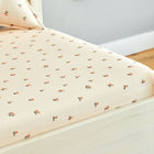 Ein helles Holzbett mit dem KraftKids Spannbettlaken Musselin Kirschen aus 100% Baumwolle, zart pfirsichfarben mit rosa-grünem Kirschmuster. Im Hintergrund ist ein passendes Kleinkinder-Bettlaken teilweise zu sehen.