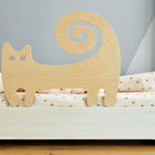 Der KraftKids Rausfallschutz aus Leimholz Katze in Form einer stilisierten Katze mit Spiralschwanz ist an einem weißen Bett mit heller Blumenbettwäsche befestigt. Eine hellgraue Wand bildet den Hintergrund.