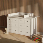 Ein KraftKids Wickelaufsatz weiß mit Blende, passend für 160 cm breite HEMNES Kommode, steht auf Holzfußboden an einer Lamellenwand. Daneben liegen ein Kuscheltier, eine Holzeisenbahn und Kinderstühle im Sonnenlicht.