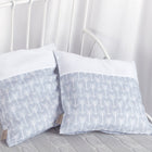 Zwei KraftKids Kissenbezüge "weiße Pfeile auf Grau" stehen dekorativ vor weißen Metallstäben eines Bettes, auf einer hellgrauen Strickdecke. Weiße Vorhänge ergänzen die gemütliche Bettwäschekollektion.