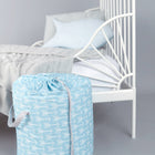 Der KraftKids Spielzeugkorb weiße Pfeile auf Blau steht auf dem Boden neben einem weißen Metallbett mit grauem Bettzeug und einem blauen Kissen und bringt Ordnung in den Raum vor grauem Hintergrund.