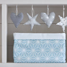 Vier Stoffornamente (zwei Herzen, zwei Sterne) in zartem Grau, Weiß und Blau schweben über dem Körbchen weiße Diamante auf Pastell Blau von KraftKids - die ideale Aufbewahrung für Ihren Wickeltisch - auf einem strahlend weißen Holzregal.