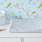 A KraftKids Bezug für Keilwickelauflage in Blau mit weißen Pfeilen liegt auf einer weißen Kommode neben passendem Korb und Schnullerkette, vor blauer Tapete mit bunten Vögeln und Libellen.
