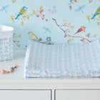 Ein KraftKids-Wickeltisch mit dem "Bezug für Keilwickelauflage kleine Dreiecke blau grau weiß", einer geometrischen blau-grau-weißen Wickelauflage, einem Aufbewahrungskorb und einer Schnullerklemme, vor einer Tapete mit bunten Vögeln und Libellen.