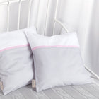 Zwei KraftKids Kissenbezüge Unigrau Karo grau mit weißem Oberteil, rosa Streifen und grau kariertem Unterteil liegen auf einer hellgrauen Strickdecke auf einem weißen Metallbett mit weißen Vorhängen im Hintergrund.