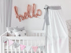 Ein weißes Babybett mit sternförmigen Kissen, einem Betthimmel Unigrau von KraftKids aus 100% Baumwolle und pastellfarbenen Wimpeln. Über der Krippe buchstabieren roségoldene Luftballons "Hallo" auf eine weiße Wand.