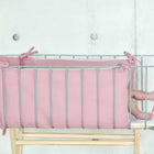 Ein Metallbettchen mit der KraftKids Bettumrandung Nestchen Musselin rosa steht vor einer hellgrauen Wand. Im Inneren sind nur die Beine und Füße des Babys zu sehen, verdeckt von der weichen Bettumrandung und dem Geländer.