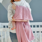 Eine Frau hält ein in die KraftKids Babydecke Musselin rosa eingewickeltes Baby vor einem weißen Kinderbett, sie trägt ein helles Hemd und Jeans. Der Raum hat eine geometrische graue und weiße Wand im Hintergrund.