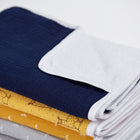 Ein Stapel gefalteter Babydecken in Marineblau, Weiß, Gelb mit Hühneraufdruck und Grau mit kleinen weißen Mustern liegt ordentlich neben der KraftKids Wickelunterlage Musselin dunkelblau auf einer weißen Fläche.