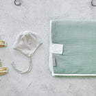 KraftKids Reisewickelunterlage Musselin mint, Babysandalen und eine gestreifte Mütze mit Kinnriemen sind ordentlich auf einer hellgrauen, strukturierten Oberfläche angeordnet.