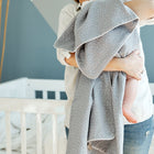 Eine Person hält ein in die KraftKids Babydecke Musselin grau Punkte eingewickeltes Baby, das neben einem weißen Kinderbett in einem schwach beleuchteten Kinderzimmer steht. Das Gesicht des Erwachsenen ist nicht zu sehen.