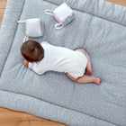 Ein Baby im weißen Strampler liegt auf der KraftKids Krabbeldecke Musselin grau Pusteblumen, die mit kuscheligem Polyestervlies gefüllt und mit zarten weißen Pusteblumenmustern versehen ist. Zwei weiche Stoffblöcke liegen daneben, und die Matte liegt auf einem Holzboden.