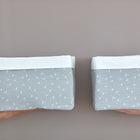 Zwei Hände halten zwei unterschiedlich große KraftKids Körbchen Musselin grau Pusteblumen mit weißem Rand vor beigem Hintergrund - ideal zur Aufbewahrung am Wickeltisch.