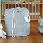 Der KraftKids Spielzeugkorb Musselin grau Pusteblumen mit weißen Griffen und einem Kordelzug sitzt auf einem Holzboden neben einem Plüschhasen und passenden Kissen und sorgt für Ordnung vor einem Holzbettchen.