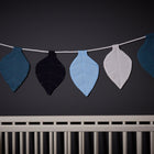 Ein weißes Kinderbett steht vor einer dunklen Wand, über der die KraftKids Wimpelkette Musselin blau dunkelblau weiß hängt - eine Girlande aus Stoffblättern in Blau, Dunkelblau und Weiß.