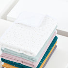 Ein ordentlich gestapelter Stapel weicher, pastellfarbener Babydecken mit kleinen Tupfen liegt neben der KraftKids Wickelunterlage Musselin goldene Punkte auf Weiß, alles auf einer weißen Fläche mit einem minimalen, hellen Hintergrund angeordnet.