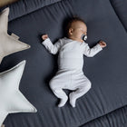 Ein Baby mit einem Schnuller schläft auf einer KraftKids Krabbeldecke Musselin goldene Punkte auf Grau, die weiß gekleidet ist. Zwei sternförmige Kissen, ein beiges und ein weißes, liegen daneben auf dem Holzboden. Die gepolsterte Matte ist sanft mit Polyestervlies gefüllt.