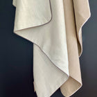 Zwei beige und hellbraune Babydecken Leinen Natur Braun von KraftKids mit sichtbarer Struktur hängen drapiert vor einem dunklen Hintergrund. Sie haben sauber verarbeitete Kanten und überlappen sich leicht, wodurch ein gemütlicher, eleganter Look entsteht.