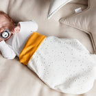 Ein Neugeborenes schläft in einem beigen Bett, eingewickelt in den KraftKids Strampelsack Frühling Sommer Musselin mit goldenen Punkten auf Weiß. In der Nähe liegen weiche, sternförmige Kissen, und das Baby hält einen Schnuller mit einem Panda-Gesicht in der Hand.