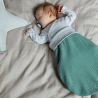 Ein Baby schläft friedlich auf einem beigen Bettchen, trägt einen grünen KraftKids Strampelsack Frühling Sommer Musselin nilblau und weißes Oberteil, mit einem Schnuller. Neben dem Baby liegen ein weiches sternförmiges Kissen und quadratische Kissen.