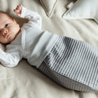 Ein Baby ruht sich auf einem cremefarbenen Bett aus. Es trägt einen grauen KraftKids Strampelsack Frühling Sommer Waffel Piqué und ein weißes Oberteil. Die Arme des Babys sind erhoben, im Hintergrund ist ein Sternenkissen zu sehen.