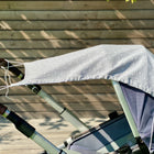 Ein Sonnensegel Musselin grau Pusteblumen von KraftKids ist über das Verdeck eines blauen Kinderwagens drapiert und bietet zusätzlichen Sonnenschutz. Der Kinderwagen steht im Freien auf einer Holzterrasse und das Sonnenlicht wirft Schatten auf die Bretterwand dahinter.