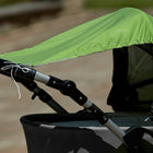 Nahaufnahme eines Kinderwagens im Freien an einem sonnigen Tag, mit dem KraftKids Sonnensegel weiße Punkte auf Grün, das mit weißen Schnüren für zusätzliche Sicherheit gesichert ist.