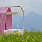 Eine KraftKids Bettbaldachin Musselin rosa hängt elegant an einem hölzernen Hausbett mit Vorhang und Schleifen, eingebettet in ein grasbewachsenes Wildblumenfeld mit Bergen und Wolken, das eine bezaubernde Kinderzimmeratmosphäre schafft.