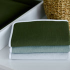 Drei ordentlich gefaltete Handtücher in Dunkelgrün, Hellblau und Weiß stapeln sich auf einer weißen Fläche neben einem geflochtenen Korb, einem zusätzlichen grünen Handtuch und der KraftKids Wickelunterlage Doppelkrepp Grün Herbstgrün.