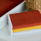 Drei ordentlich gefaltete Textilien in Brandorange, Gelb und Weiß sind auf einer weißen Fläche neben einem geflochtenen Korb und der KraftKids Wickelunterlage Doppelkrepp Rot Herbstrot gestapelt.