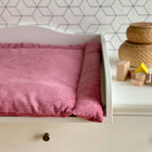 Ein weißer Wickeltisch mit einer rosa KraftKids Wickelauflage Cord Breitcord Rosa, geflochtenen Körben und Holzklötzen - darunter ein gelbes Dreieck - vor einer weißen Wand mit geometrischem Muster.