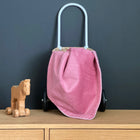 Ein kleines Holzspielzeugpferd auf Rädern steht neben einer KraftKids Babydecke Cord Breitcord Rosa Trolley Bag und einer weichen Musselin Decke, die alle auf einem hellen Holzschrank vor einem dunkelgrauen Hintergrund ausgestellt sind.