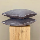 Ein KraftKids Kissenbezug Cord Breitcord Grau sitzt auf einem Holzhocker, in dem sich sein Spiegelbild spiegelt und so für Symmetrie sorgt. Weiße Paspelkanten fügen Details hinzu, während der schlichte beigefarbene Hintergrund dieses elegante Stück Bettwäschekollektion hervorhebt.