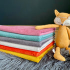 Ein Stapel von sechs gerippten, farbenfrohen Decken ruht auf einer weichen grauen Oberfläche neben einer Plüschfuchs-Puppe - eine gemütliche Einrichtung, die sich ideal mit der KraftKids Wickelunterlage Cord Breitcord Rosa kombinieren lässt.