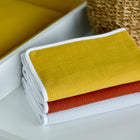 Ein ordentlich gefalteter Stapel strukturierter Handtücher in Gelb, Rost und Weiß liegt neben einem geflochtenen Korb, der die KraftKids Wickelunterlage Doppelkrepp Gelb Senf beim Windelwechseln perfekt ergänzt.