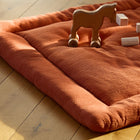 Eine kuschelige Krabbeldecke Doppelkrepp Rot Herbstrot von KraftKids liegt auf einem Holzboden. In der Nähe stehen ein Spielzeugpferd auf Rädern und Holzklötze, während das Sonnenlicht die weiche Polyestervlies-Füllung hervorhebt.