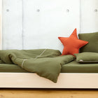 Ein minimalistisches Bett mit dem Bettwäscheset Doppelkrepp Grün Herbstgrün von KraftKids, einem orangefarbenen Sternkissen und einem grünen Kissen auf einem hellen Holzrahmen vor einer grauen Betonwand. Der helle Holzboden unterstreicht das erholsame Schlaf-Ambiente.