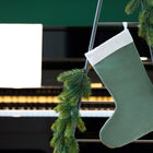 Eine Weihnachtssocke Doppelkrepp Grün Jade von KraftKids hängt an einer Leiter mit Tannengirlande, während im Hintergrund ein sanft verschwommenes Klavier und Notenblätter erscheinen.