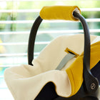 Eine Nahaufnahme einer modernen Babyschale mit gelbem Griff und Zierleiste, die mit der KraftKids Decke für Babyschale Winter Doppelkrepp Gelb Senf versehen ist, drinnen in der Nähe eines Fensters mit weißen Jalousien und Grünpflanzen draußen.