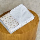 Ein gefaltetes weißes Handtuch mit einem dezenten braunen Blattmuster liegt auf einem runden Holzhocker neben der KraftKids Wickelunterlage Musselin Zweige braun, vor einem beigefarbenen, strukturierten Hintergrund.